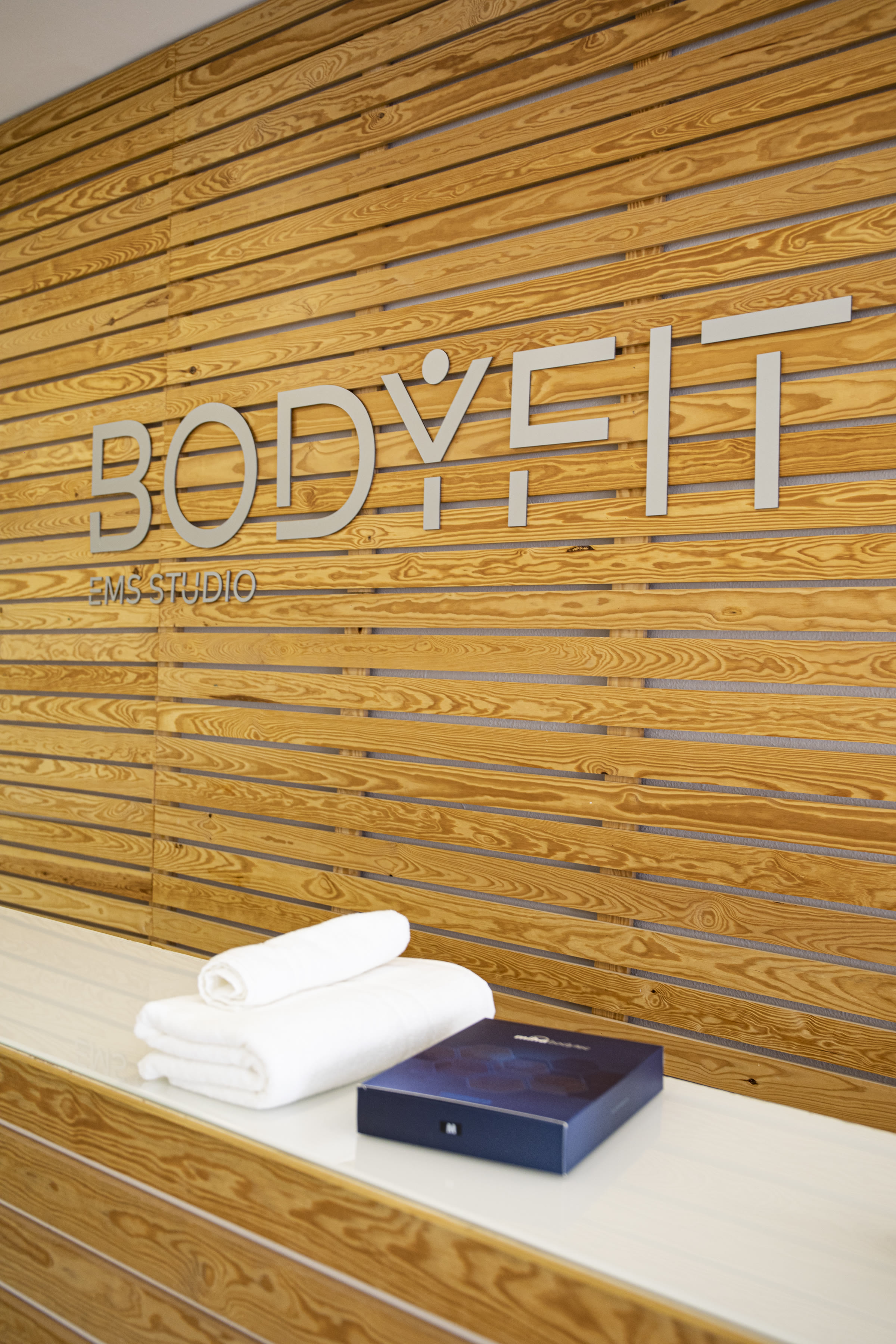 Receção do BODYFIT EMS Studio em Campo de Ourique, com logo BODYFIT em letras metálicas sobre parede de madeira clara e toalhas brancas dobradas.