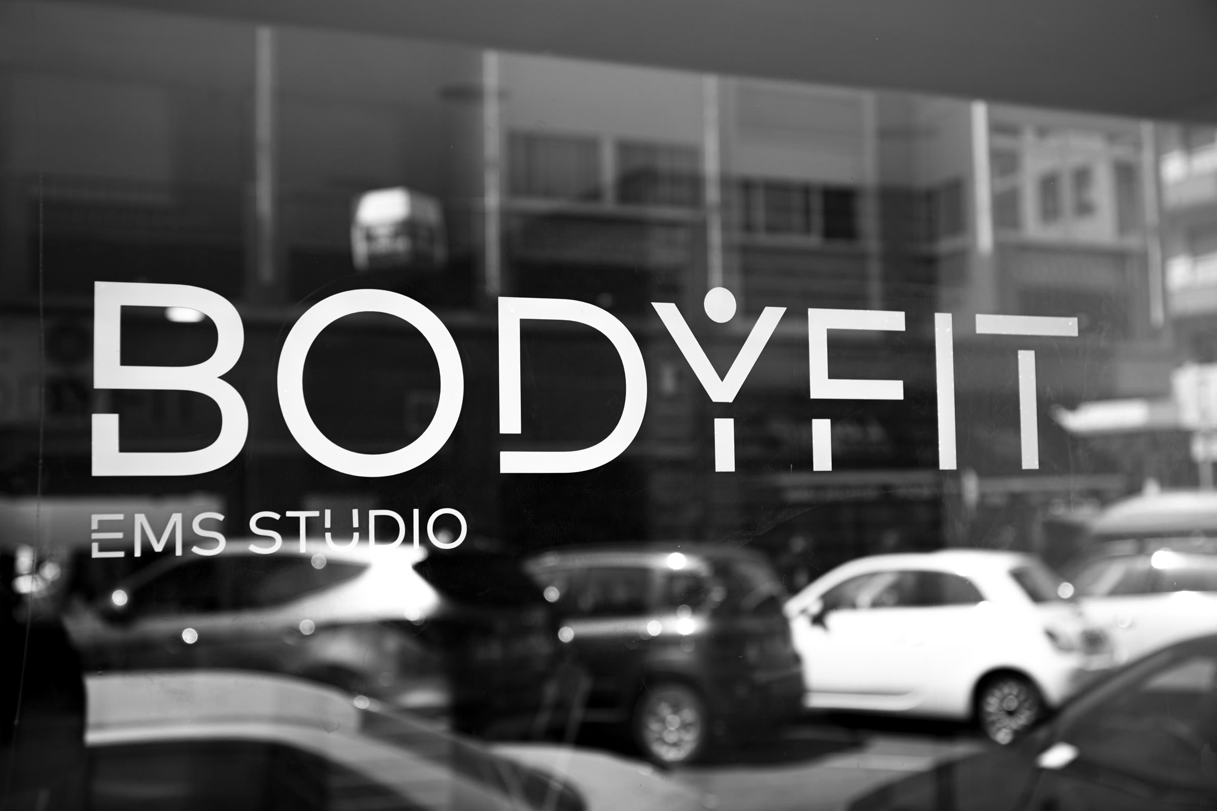 Vitrine exterior do BODYFIT EMS Studio em Campo de Ourique, Lisboa, com o logo BODYFIT em vinil branco e o reflexo da rua a preto e branco.